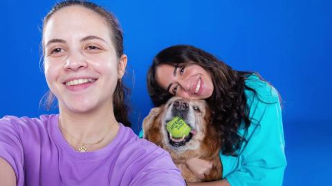 Two girls are smiling and one of them is holding a dog that has a tennis ball in his mouth