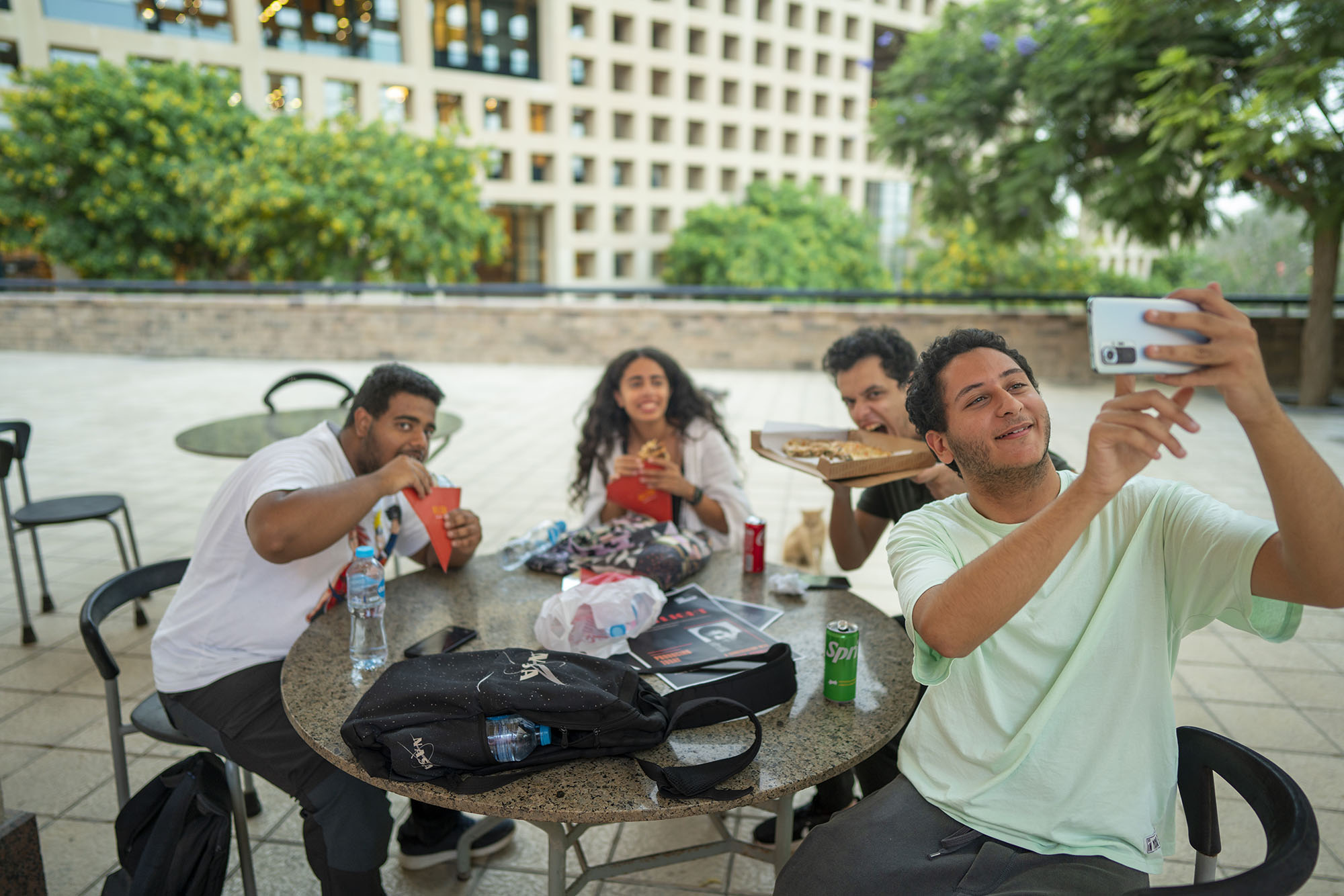 Group of girls and boys sitting outdoors taking a selfie while eating