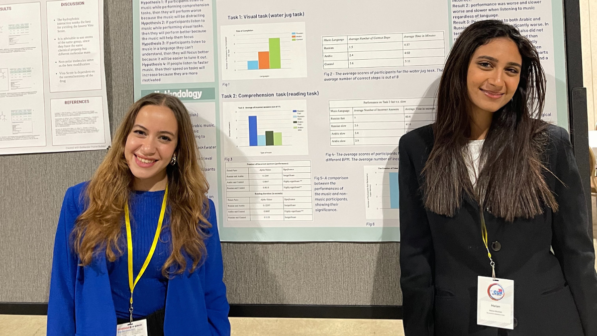 Two women, one in a blue jacket and another in a black jacket, smile in from of a large scientific poster