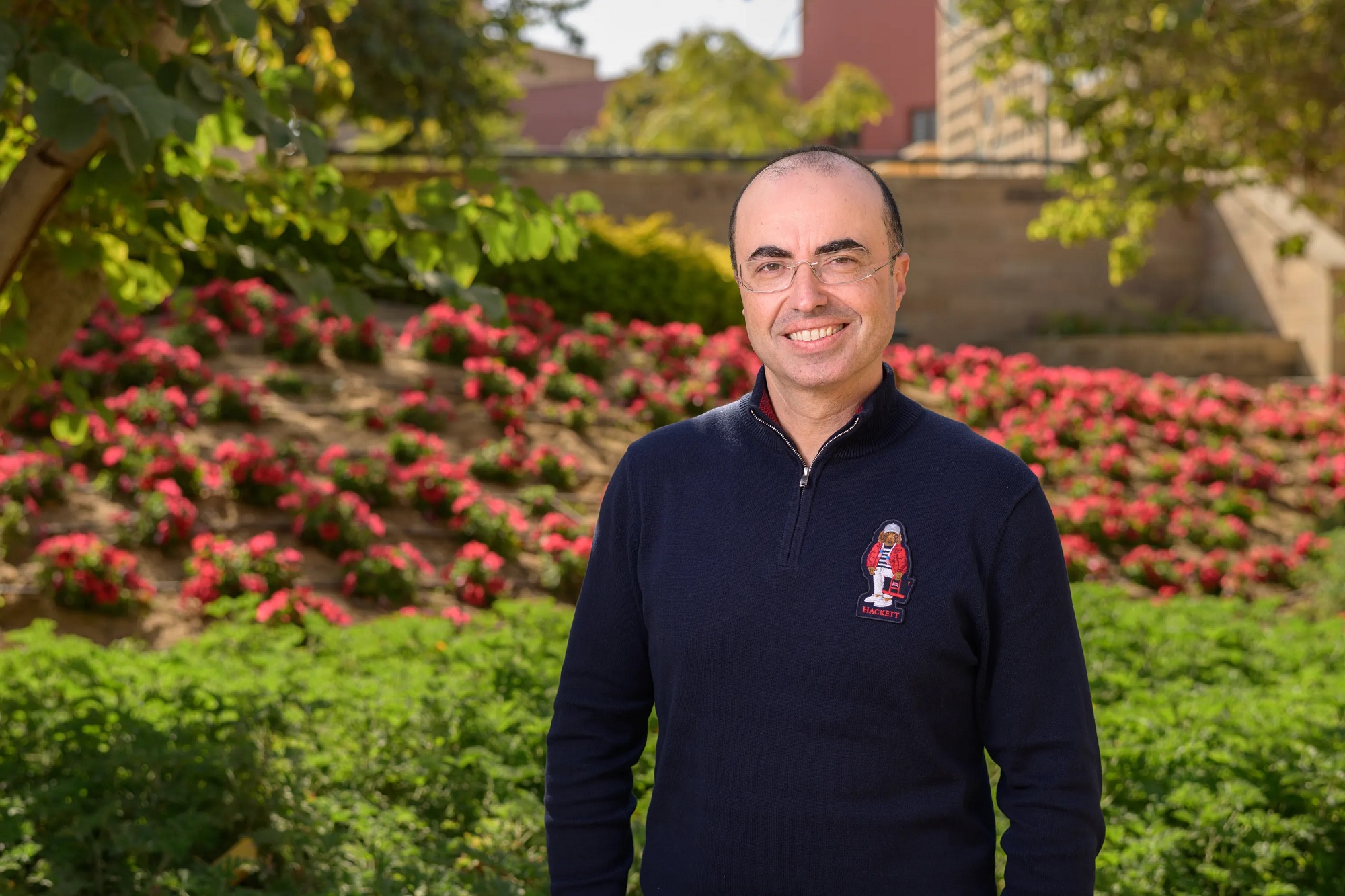 A man is standing in a garden and he is smiling. He is wearing glasses