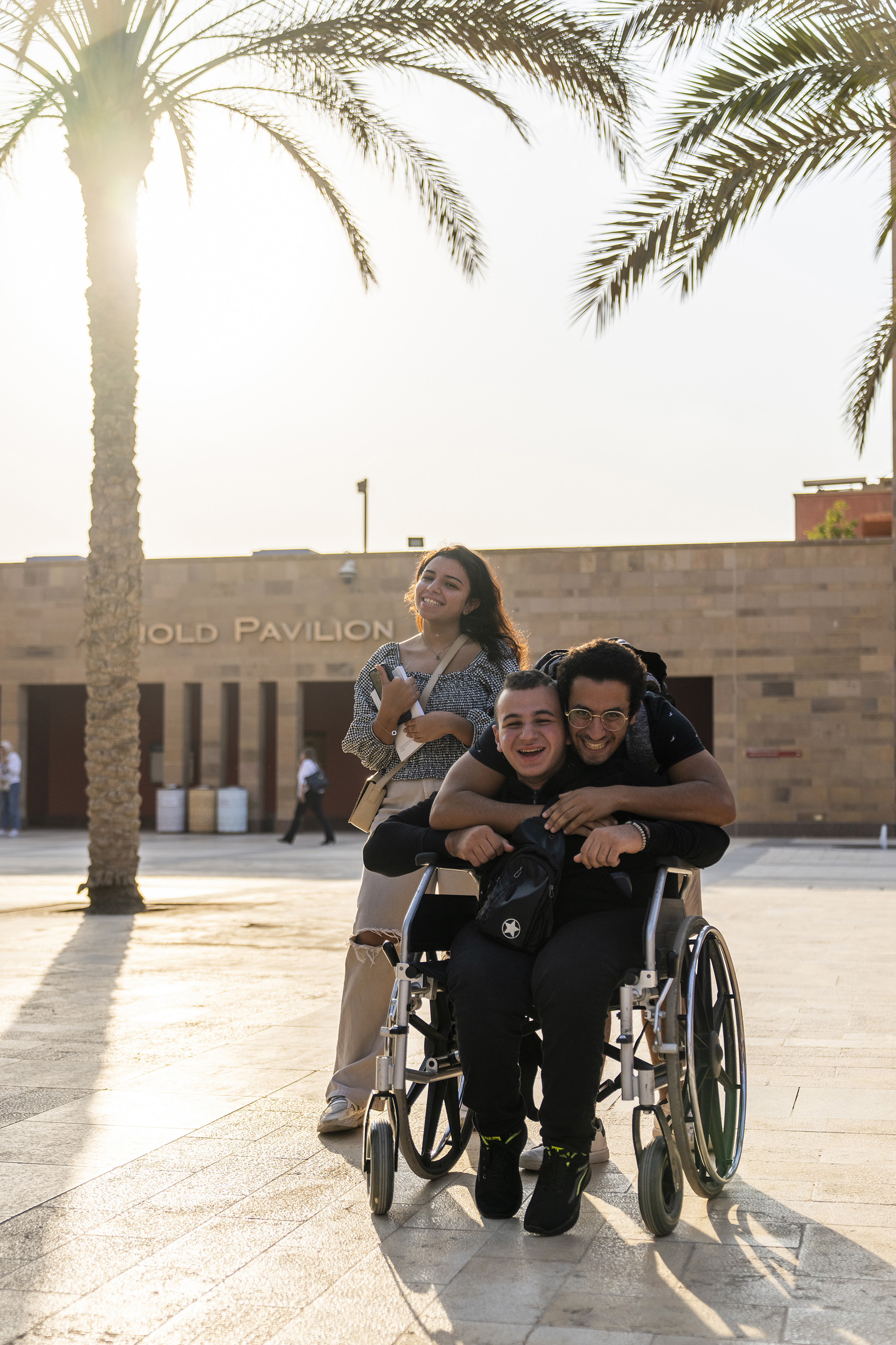 A boy sitting on a wheel chair smiling and behind him a boy and a girl standing