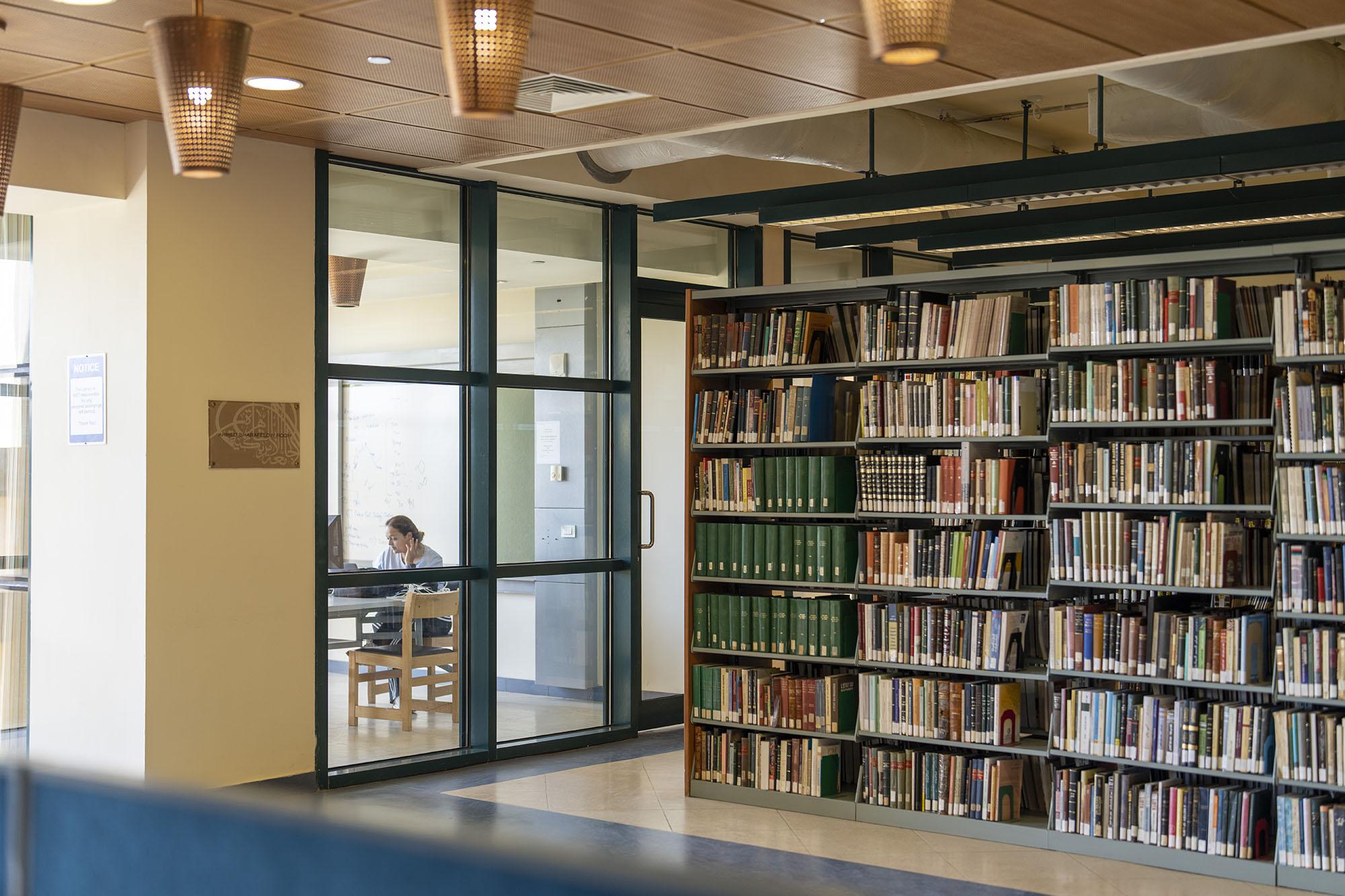 Female student studying in a glass room in the library next to book shelves