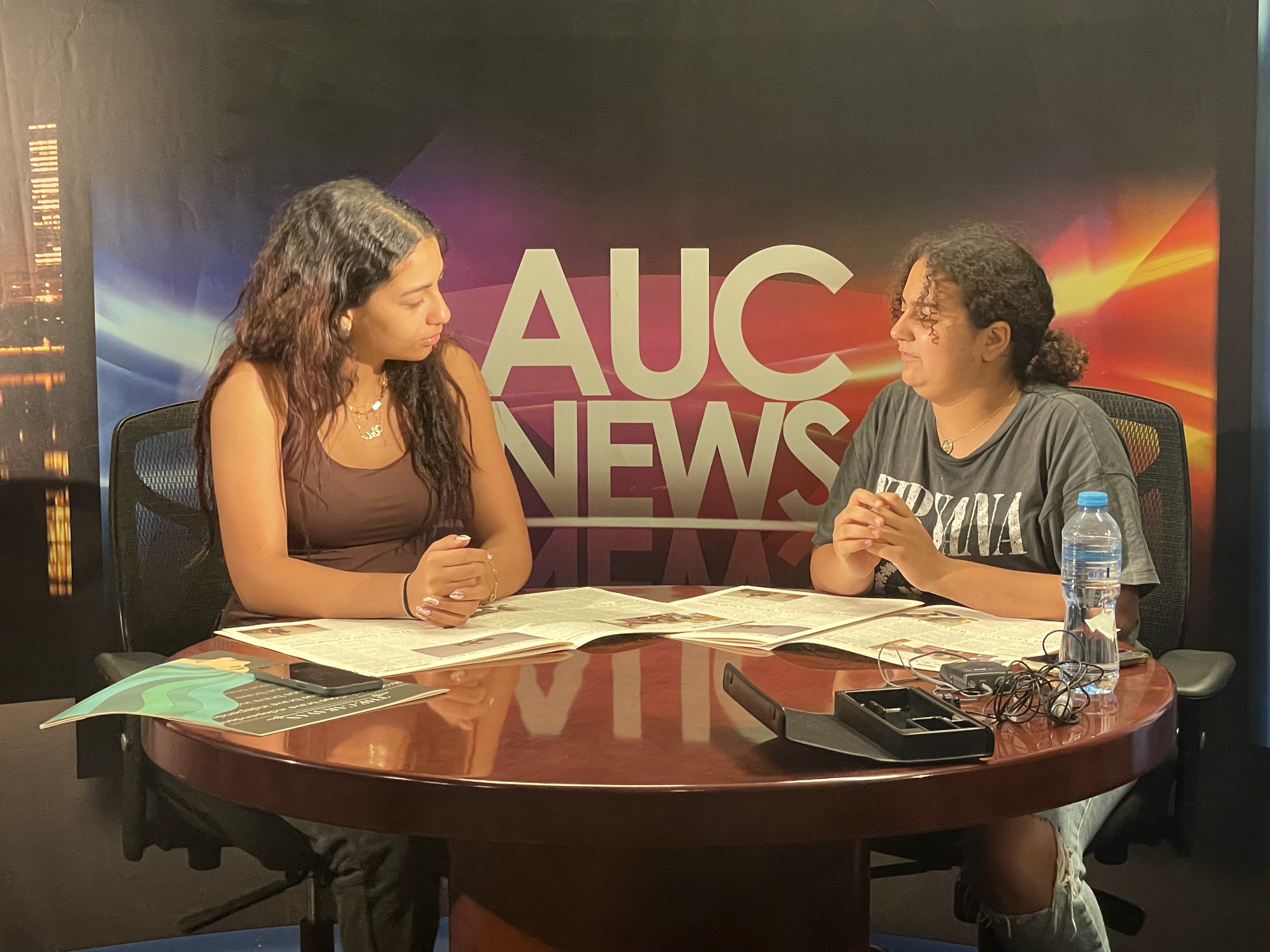 2 students sitting on a media interview table with AUC News banner behind them