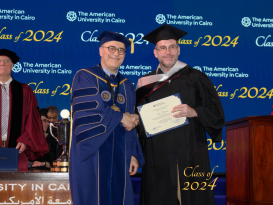 A professor in a cap and gown stands holding a certificate shaking hands with the President of AUC