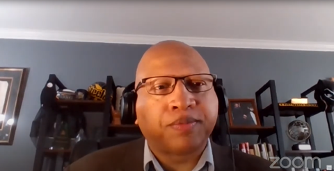 A man is wearing glasses and a brown jacket and white shirt, there is a shelf, it has a globe and photo frame. Text: Zoom