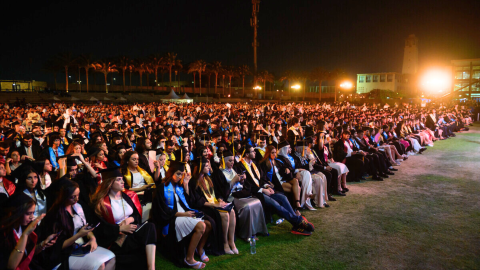 A large group of graduating students in gowns and caps sit together facing the stage