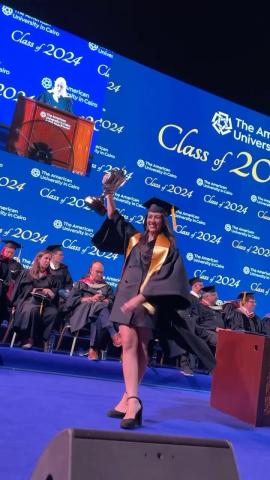 A female student wearing her cap and gown and holding a winners cup. There are people sitting behind her wearing caps and gowns. Text: The American University in Cairo