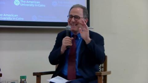 A man is wearing a suit and glasses. He is sitting on a chair and talking into a microphone