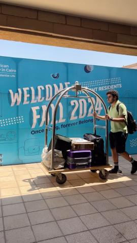 A male is pushing a luggage cart, it has bags on it. He is walking next to a wall with the text: Welcome Fall 2024. Student Residences