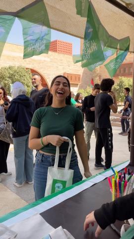 People are standing. A female is standing and laughing. She is holding a bag with the text: Mental Health Week