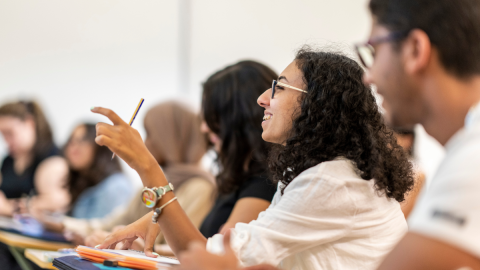 There is a male and female students sitting in class. A female student is talking and holding a pencil in her hand