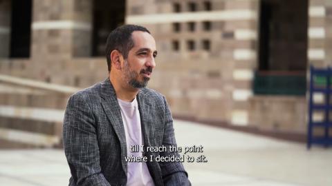 A man is talking outdoors. He is wearing a gray jacket and lilac t-shirt