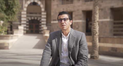 A man is talking. He is wearing glasses, a dark beige jacket and a white shirt