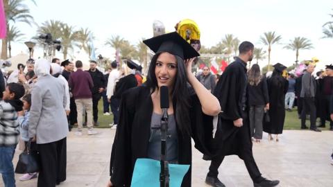 A female is wearing her cap and gown. There are people walking around her