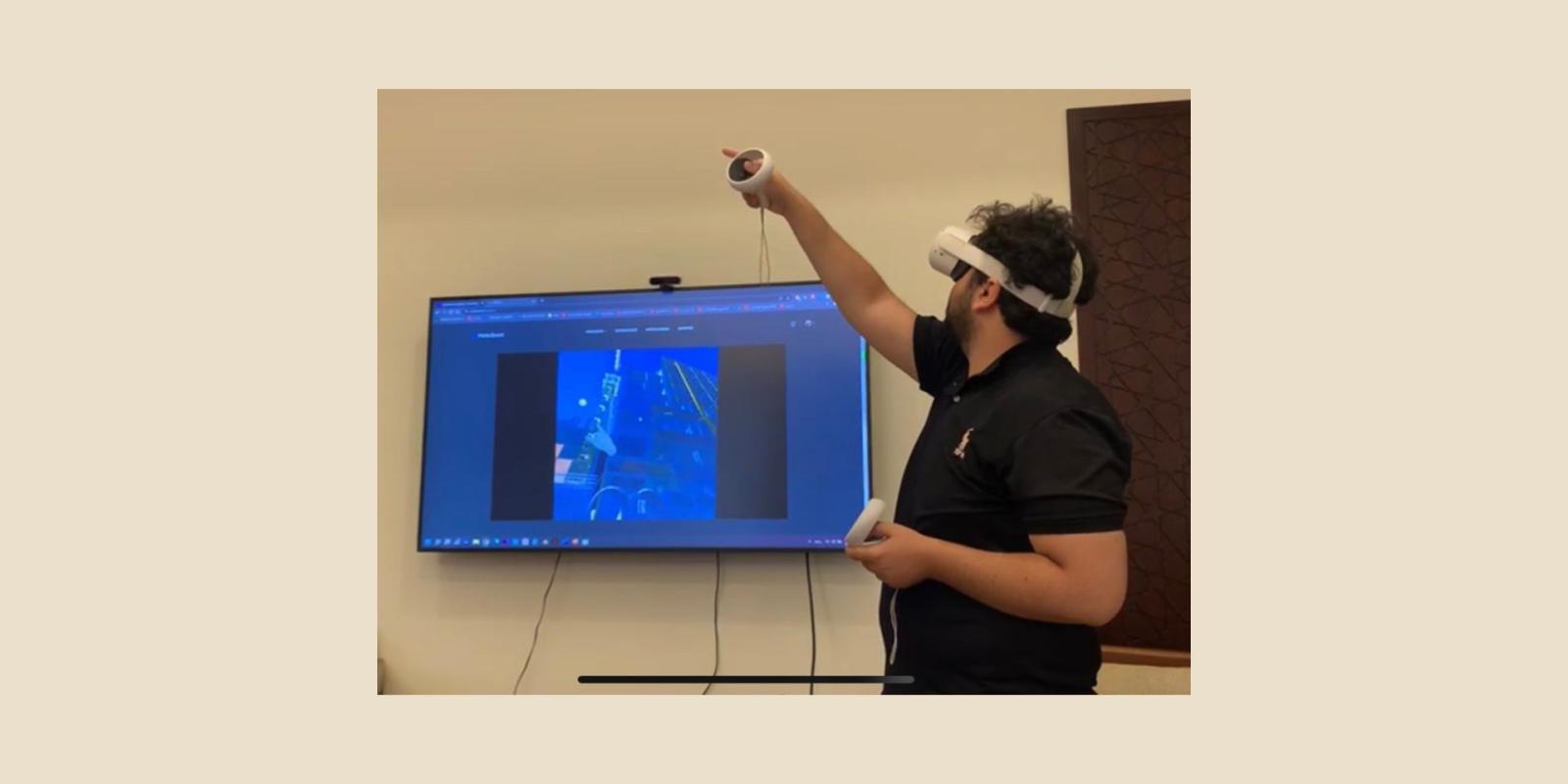 Man stands wearing a virtual reality headset and controllers while a screen behind him shows what he is seeing through the equipment