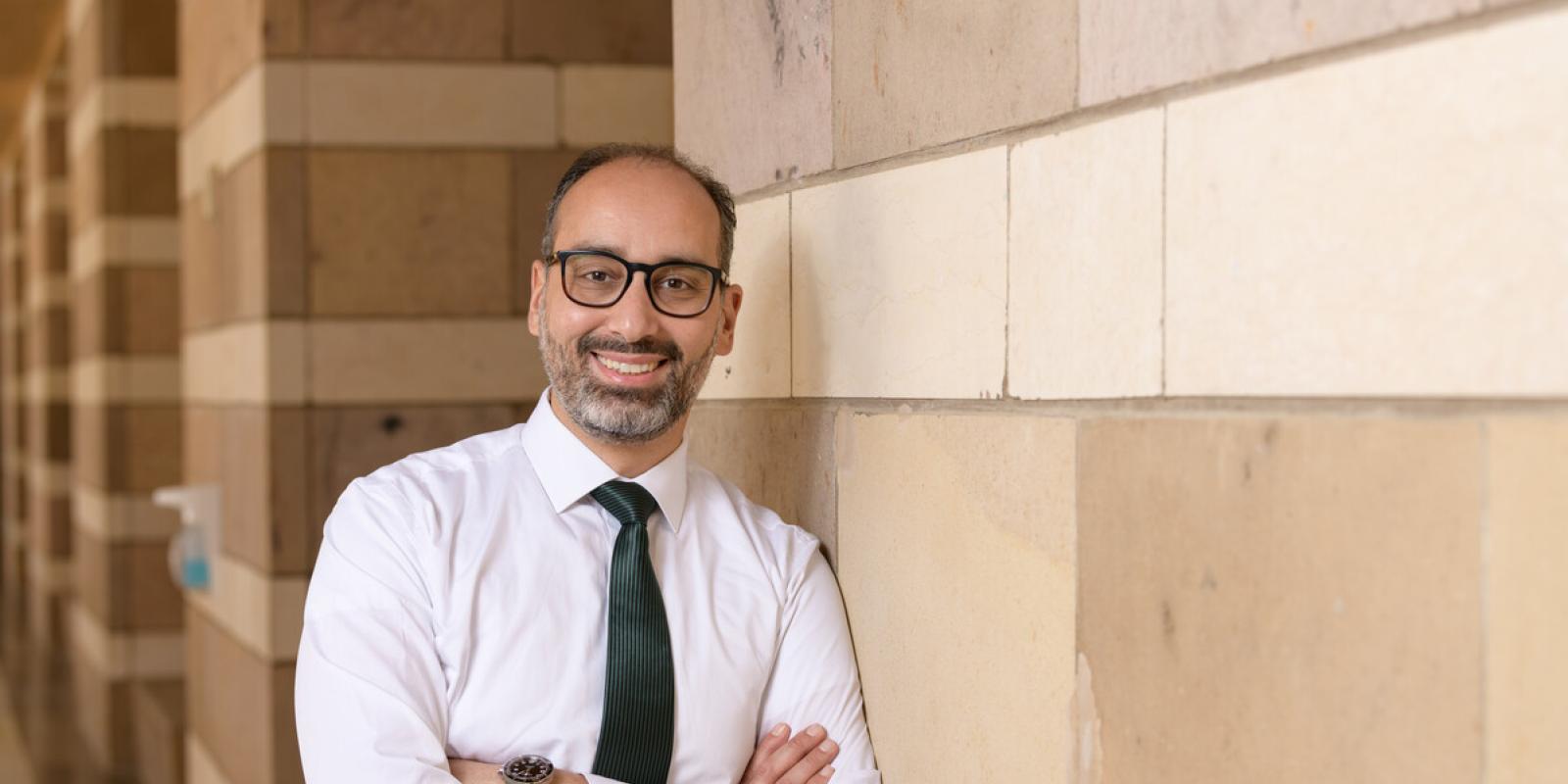 Smiling man in glasses wearing a button-up and blue tie leaning against a wall at AUC