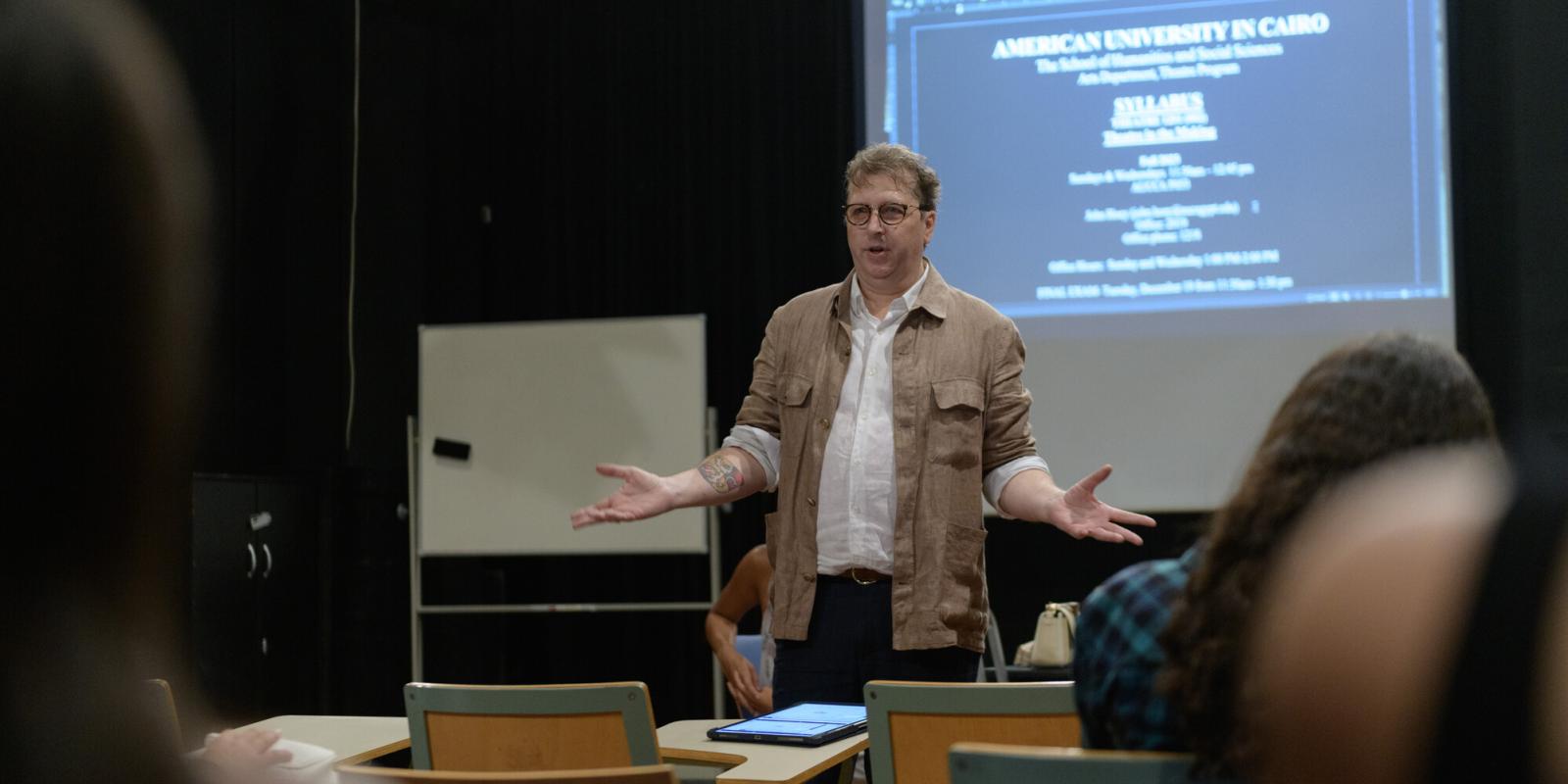 A man explaining in class with a projector behind him