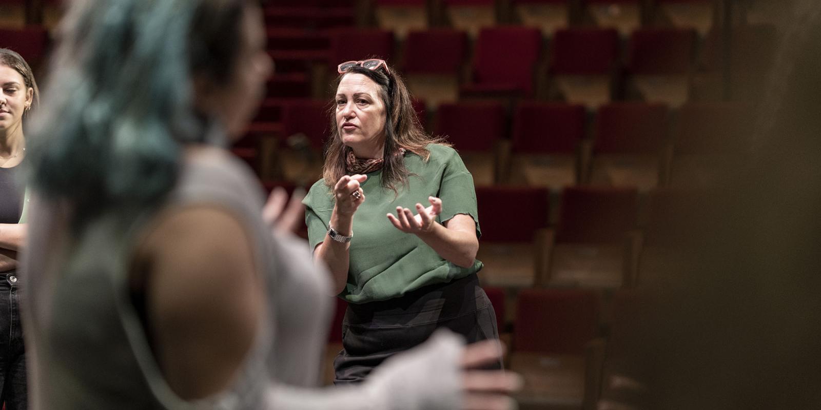 A woman in a green shirt stands in front of red theater chairs, a student in a gray shirt in the foreground