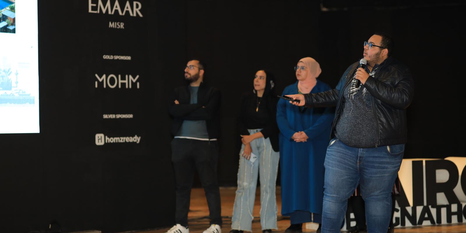 Man in a black jacket and blue jeans presents in front of a conference background