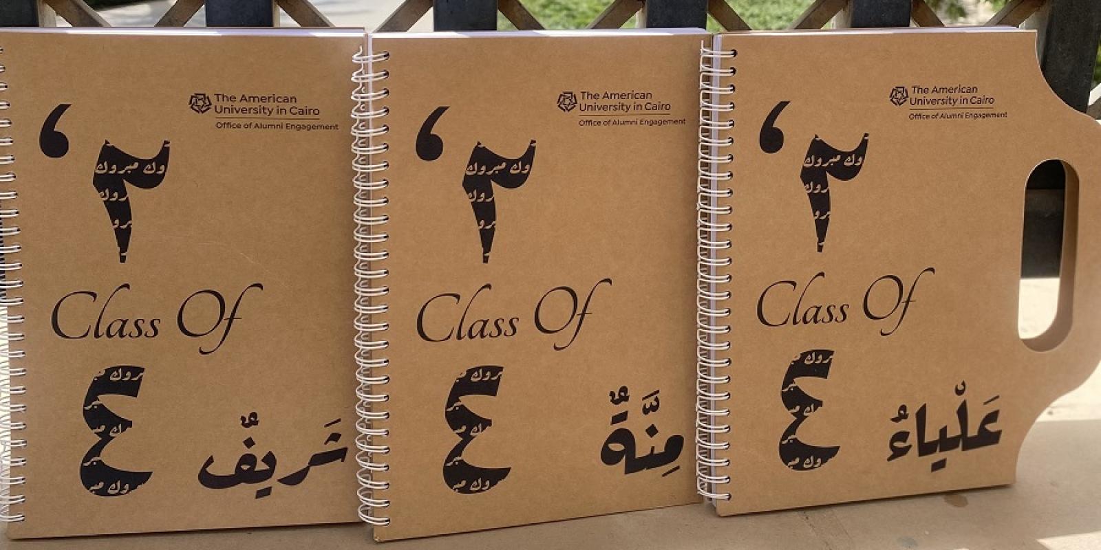 Notebooks. Text: The American University in Cairo. Office of  Alumni Engagement. Class of 24 in Arabic numbers. The names Aliaa, Menna and Sherif in Arabic