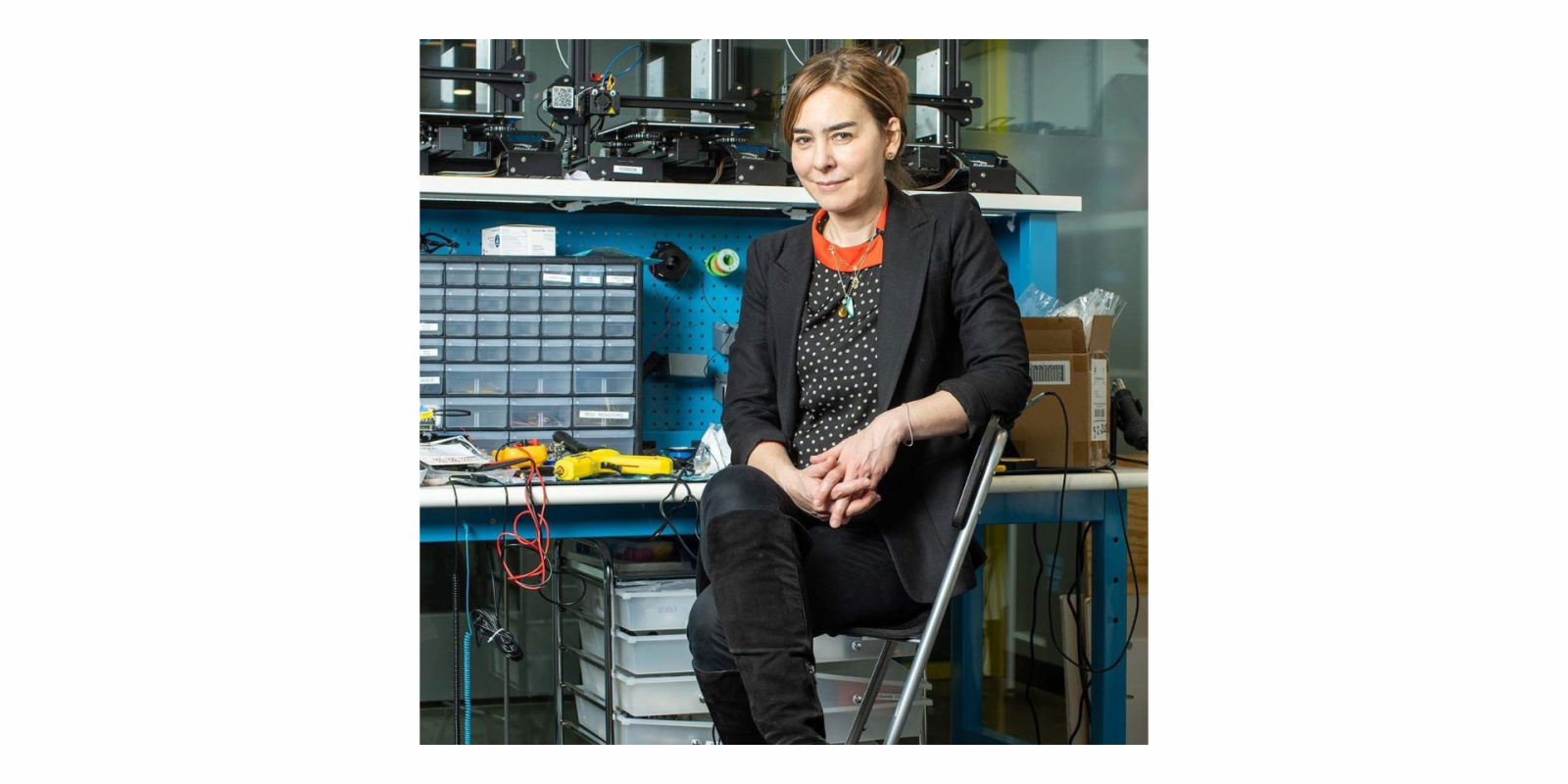Woman sits in a dark jacket inside of a workspace filled with tools and machines
