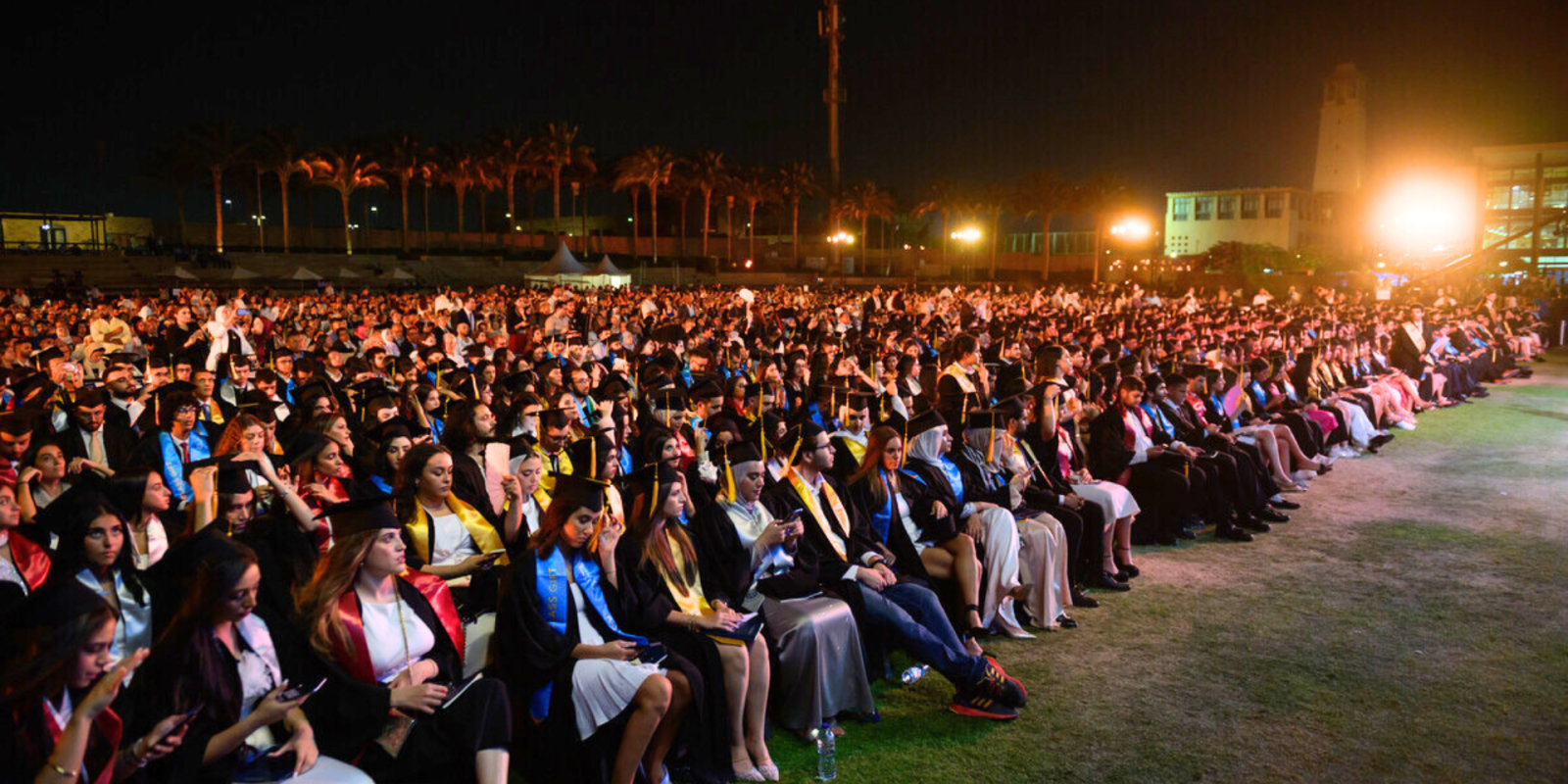 A large group of graduating students in gowns and caps sit together facing the stage