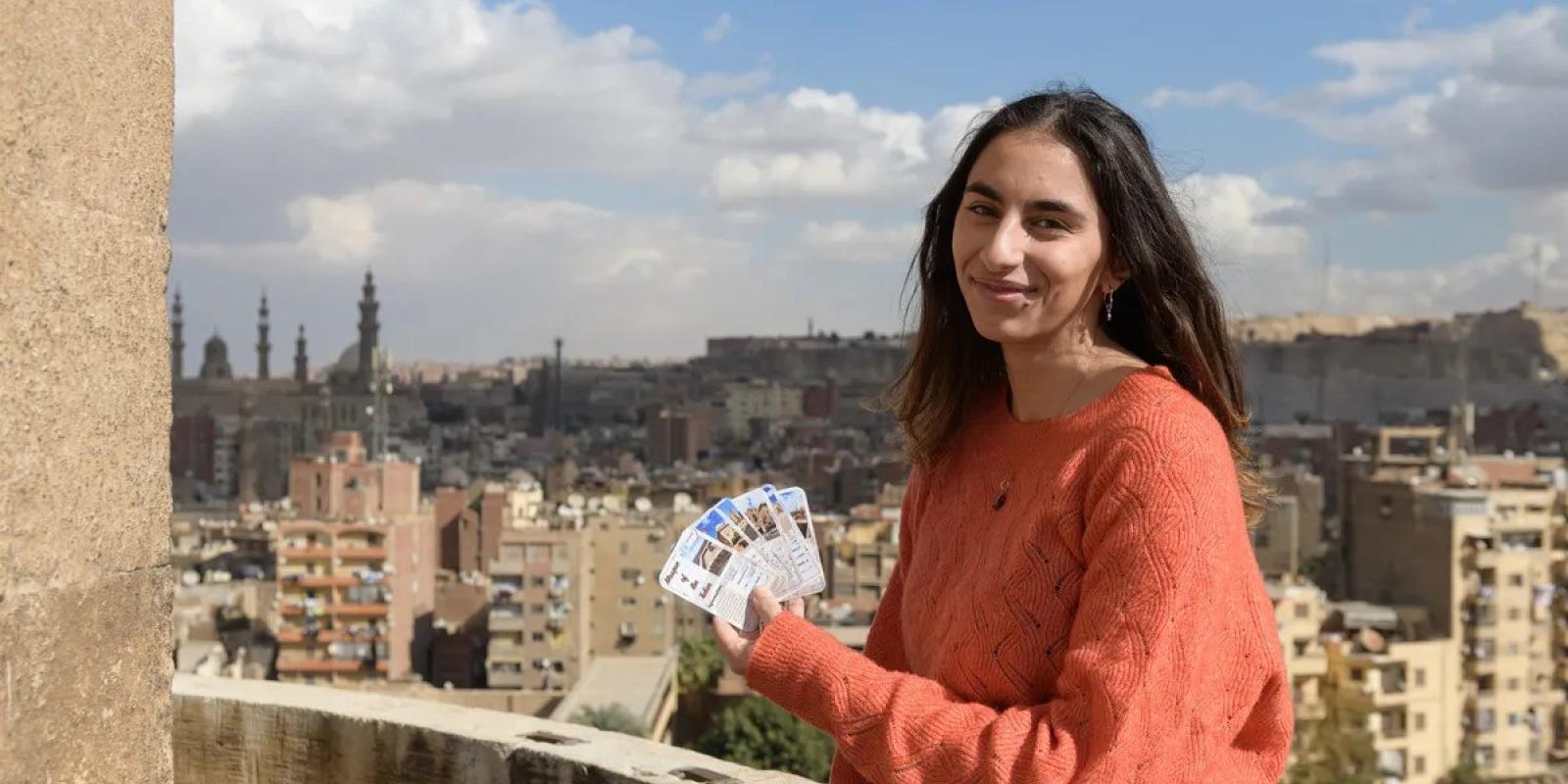 A girl is smiling, she is holding cards with pictures on them. There are buildings and mosques behind her