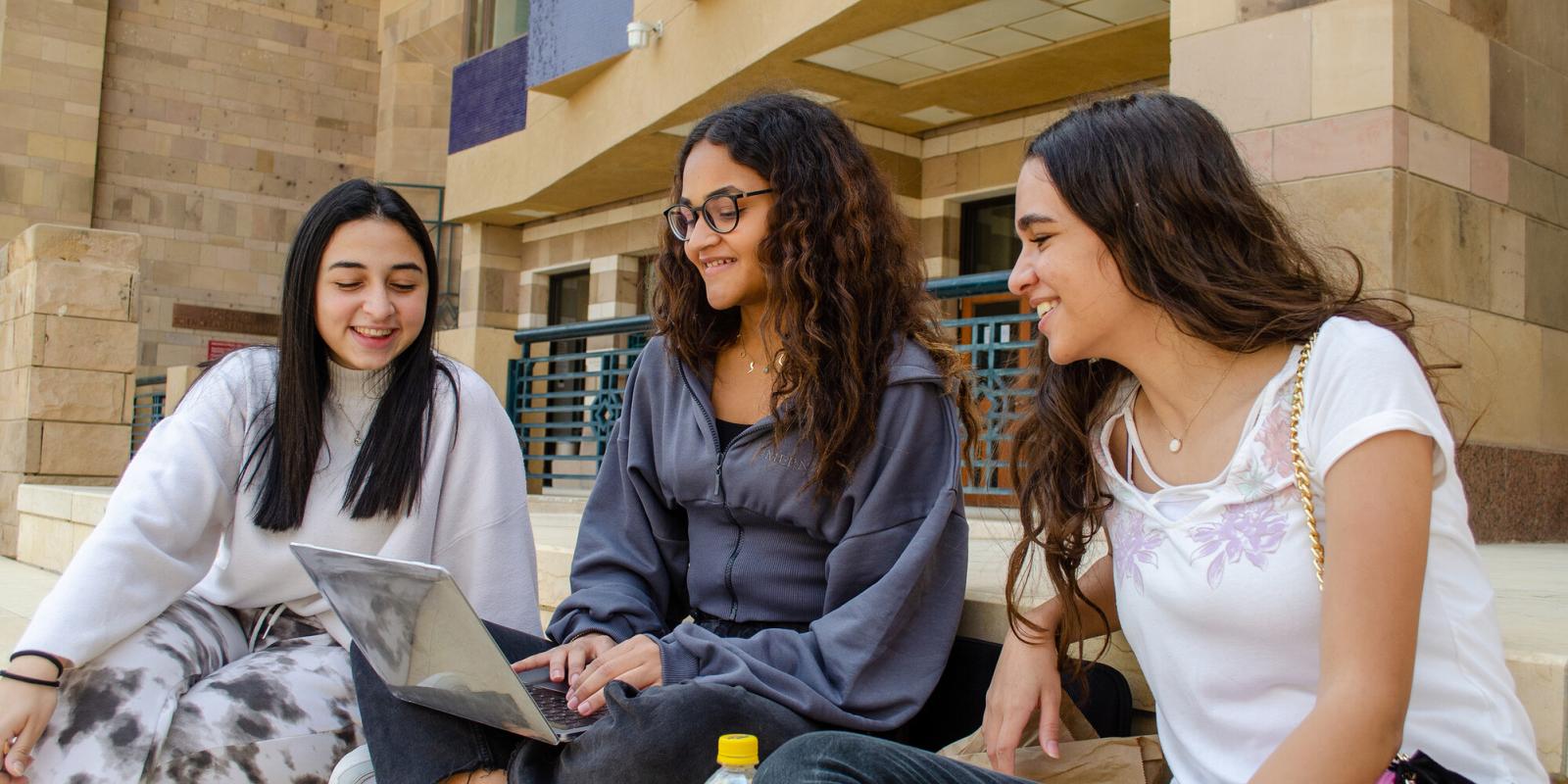 A group of three students sitting together smiling and looking a laptop