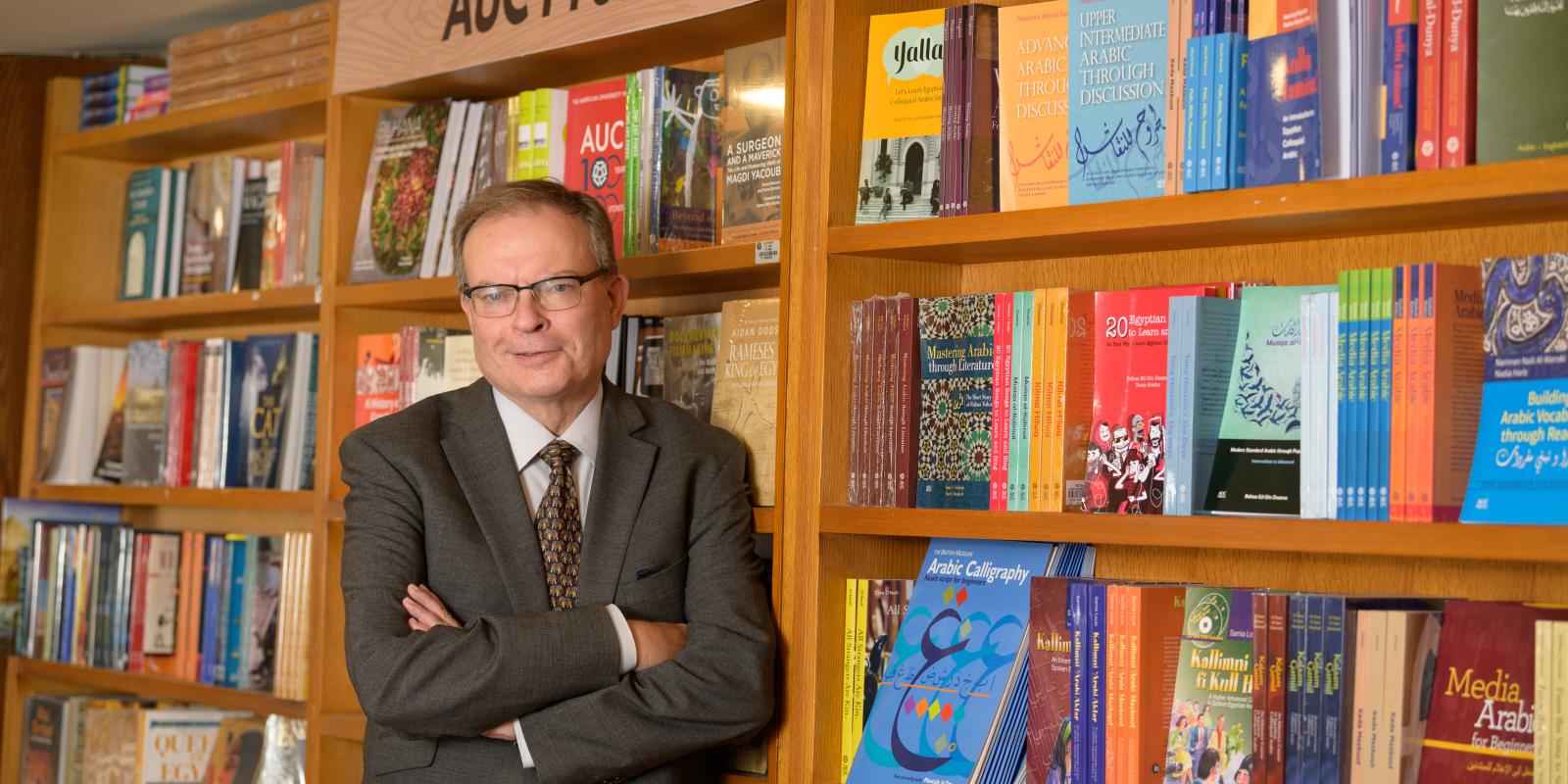 Man wearing a suit in front of bookshelf at the university bookstore, shelf section labelled "AUC Press"