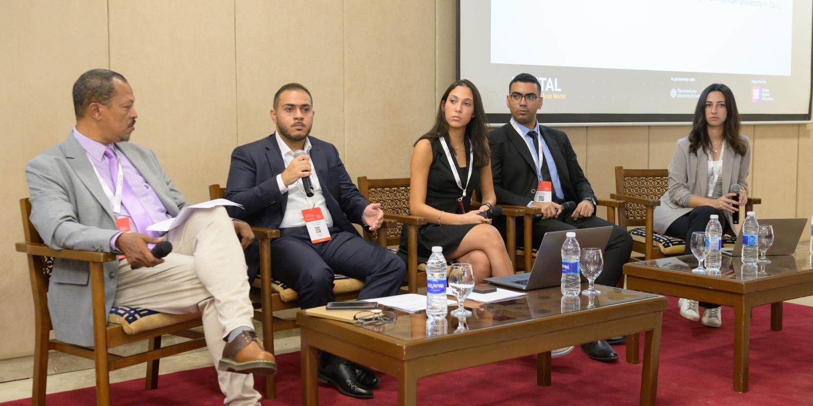 Four students and one faculty moderator sit in a row in front of a low table.