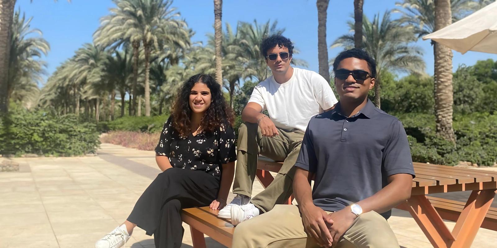 Two men and one woman sit on a picnic table in the AUC garden, palm trees in the background