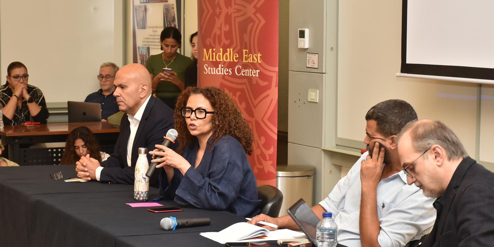 Four people sitting at a panel table. Woman speaking into a microphone.