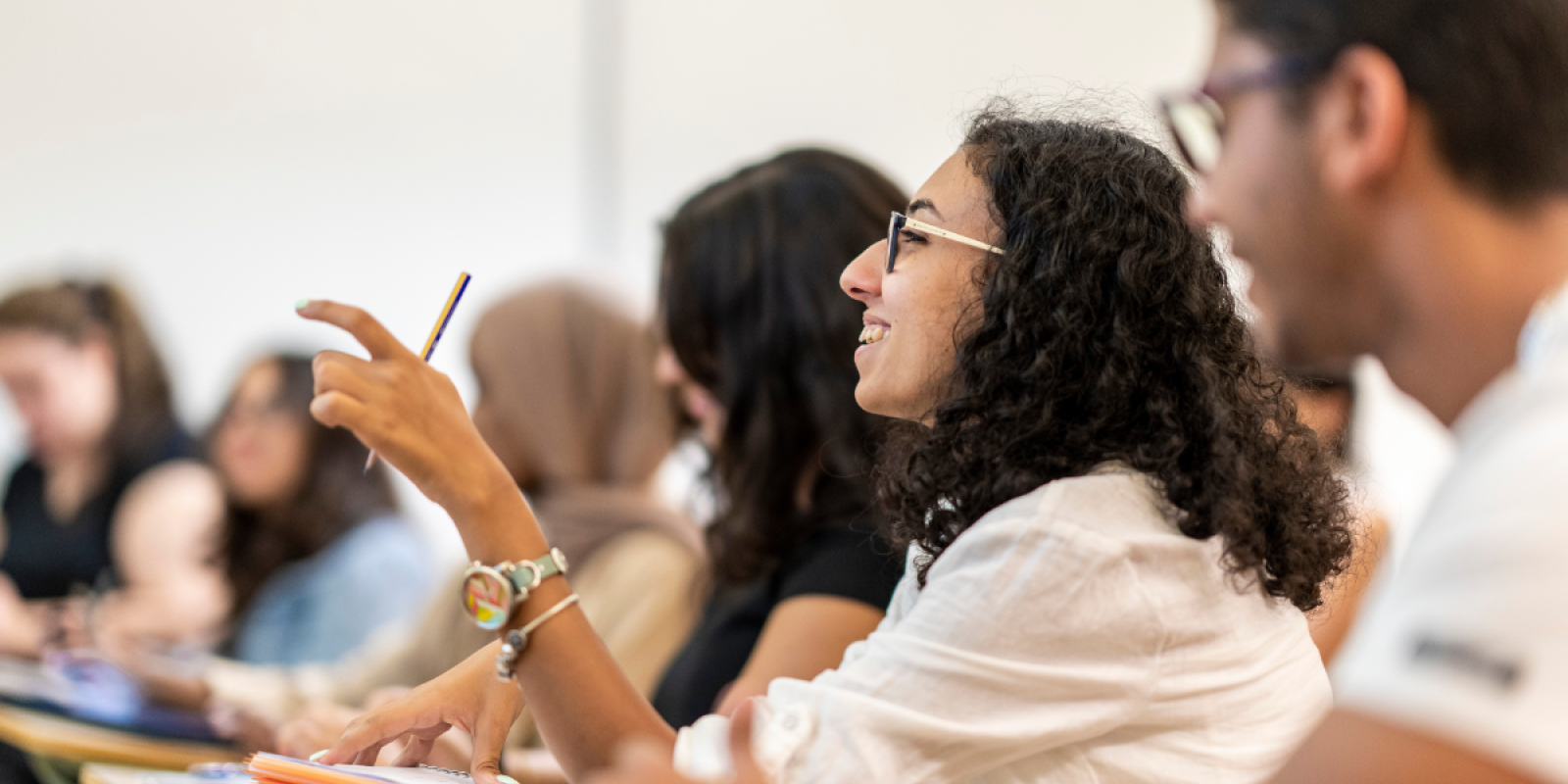 There is a male and female students sitting in class. A female student is talking and holding a pencil in her hand