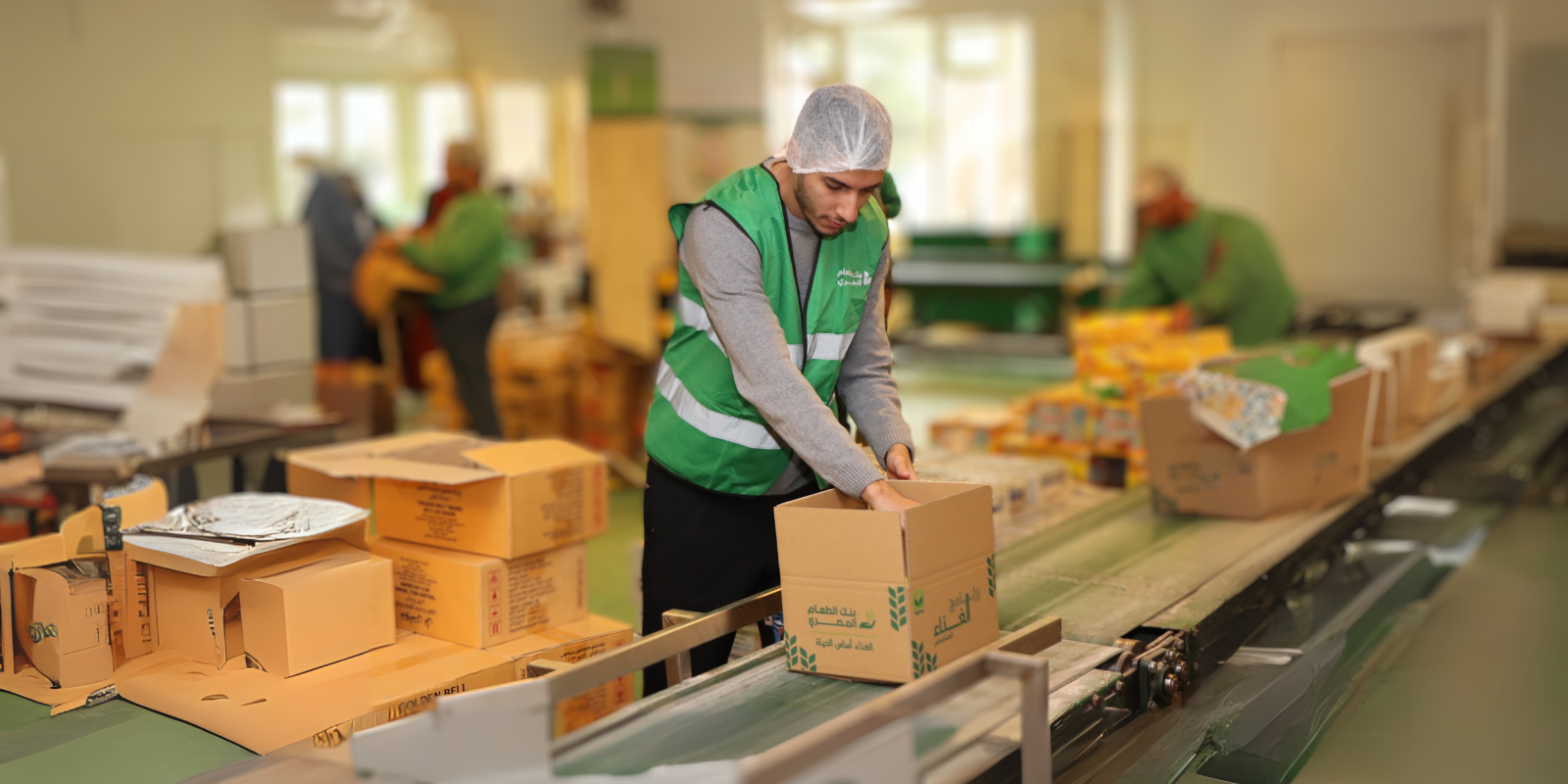 Man wearing green vest and hairnet packs a box with food supplies in a warehoues