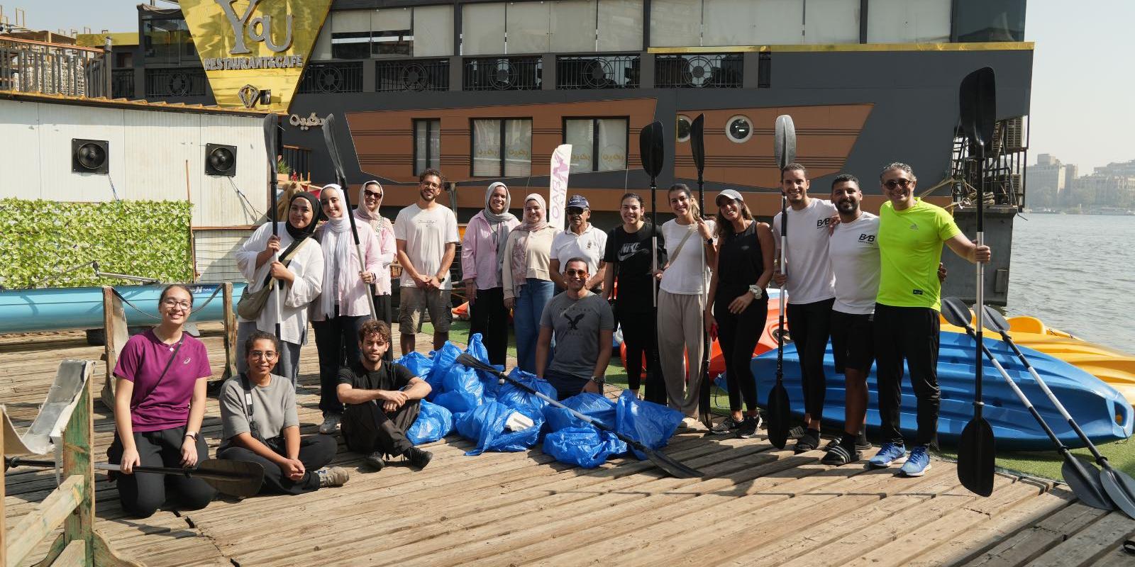 A group of students and faculty standing together on Qursaya Island with plastic bags next to them that have trash colelcted from the Nile