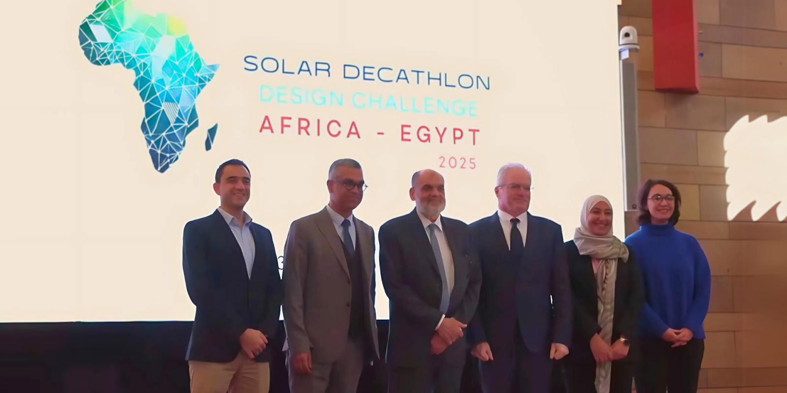 Administrators stand in front of a stage with a projection that reads "Solar Decathlon Design Challenge Africa - Egypt" with a play button