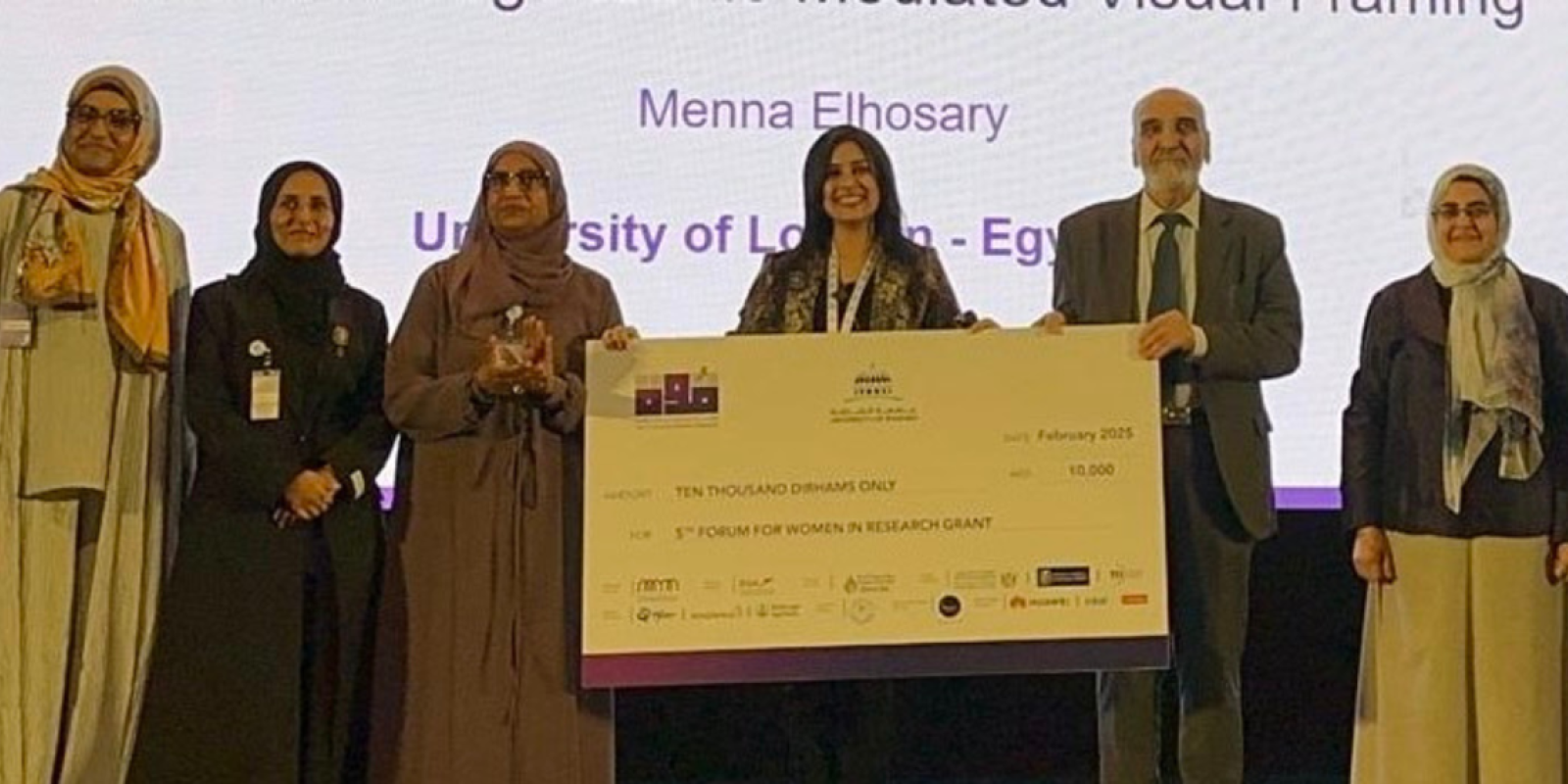 Alumna Menna El Hosary holding her winning certificate and smiling with a group of people standing next to her on stage