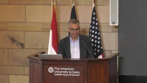 Man wearing a suit talking on a podium with 3 flags behind him