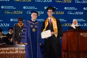 A man stands in a cap and gown in front of a commencement background holding a certificate and a golden trophy next to the AUC President.