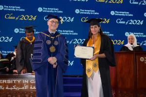 A woman stands in a cap and gown in front of a commencement background holding a certificate next to the AUC President.