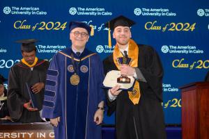 A man in a cap and gown stands next to the President of AUC holding a certificate and a golden trophy