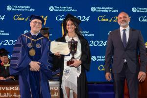 A woman stands in a cap and gown in front of a commencement background holding a certificate and a golden trophy next to the AUC President.