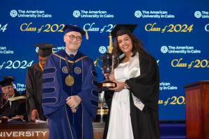 A woman in a cap and gown stands next to the President of AUC holding a certificate and a golden trophy