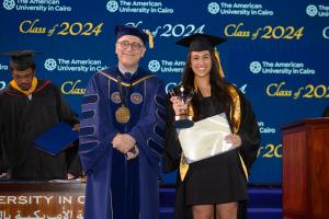 A woman in a cap and gown stands next to the President of AUC holding a certificate and a golden trophy