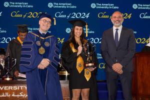 A woman in a cap and gown stands next to the President of AUC holding a certificate and a golden trophy