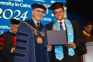 Man in a cap and gown stands next to the AUC President holding a certificate