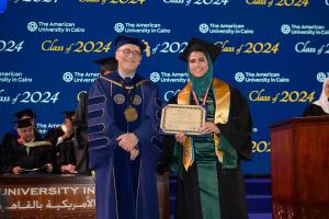 A woman stands in a cap and gown in front of a commencement background holding a certificate next to the AUC President.