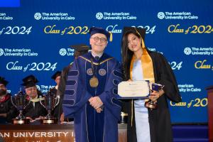 A woman in a cap and gown stands next to the President of AUC holding a certificate and a golden trophy