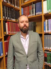 A bearded man is wearing a beige jacket and pink shirt. He is standing between library book shelves