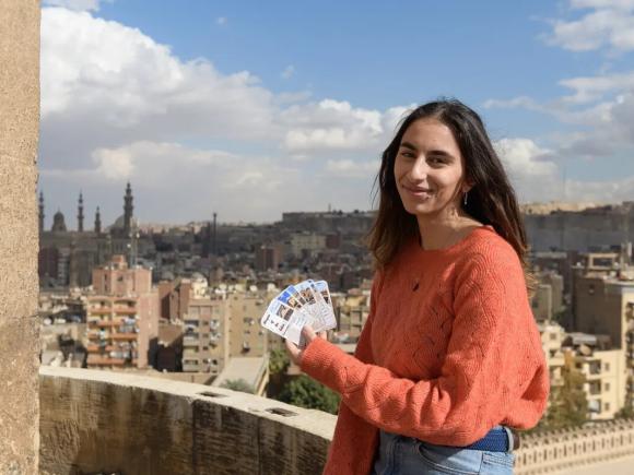 A girl is smiling, she is holding cards with pictures on them. There are buildings and mosques behind her